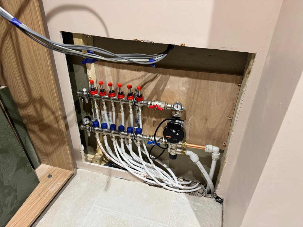 Recessed underfloor heating manifold with stainless flow and return rails, red flow meters, blue return ports, circulation pump and white flexible pipes to floor loops